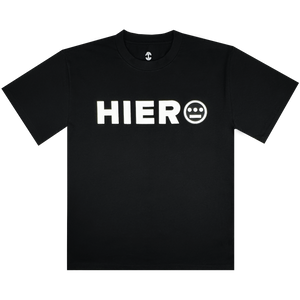 The Hiero Bold Heavy Tee by Hieroglyphics is a black T-shirt with "HIERO" in bold white letters on the chest, inspired by hip-hop culture. It features a face-like white circle with three lines and two dots, round neckline, and short sleeves against a white background.